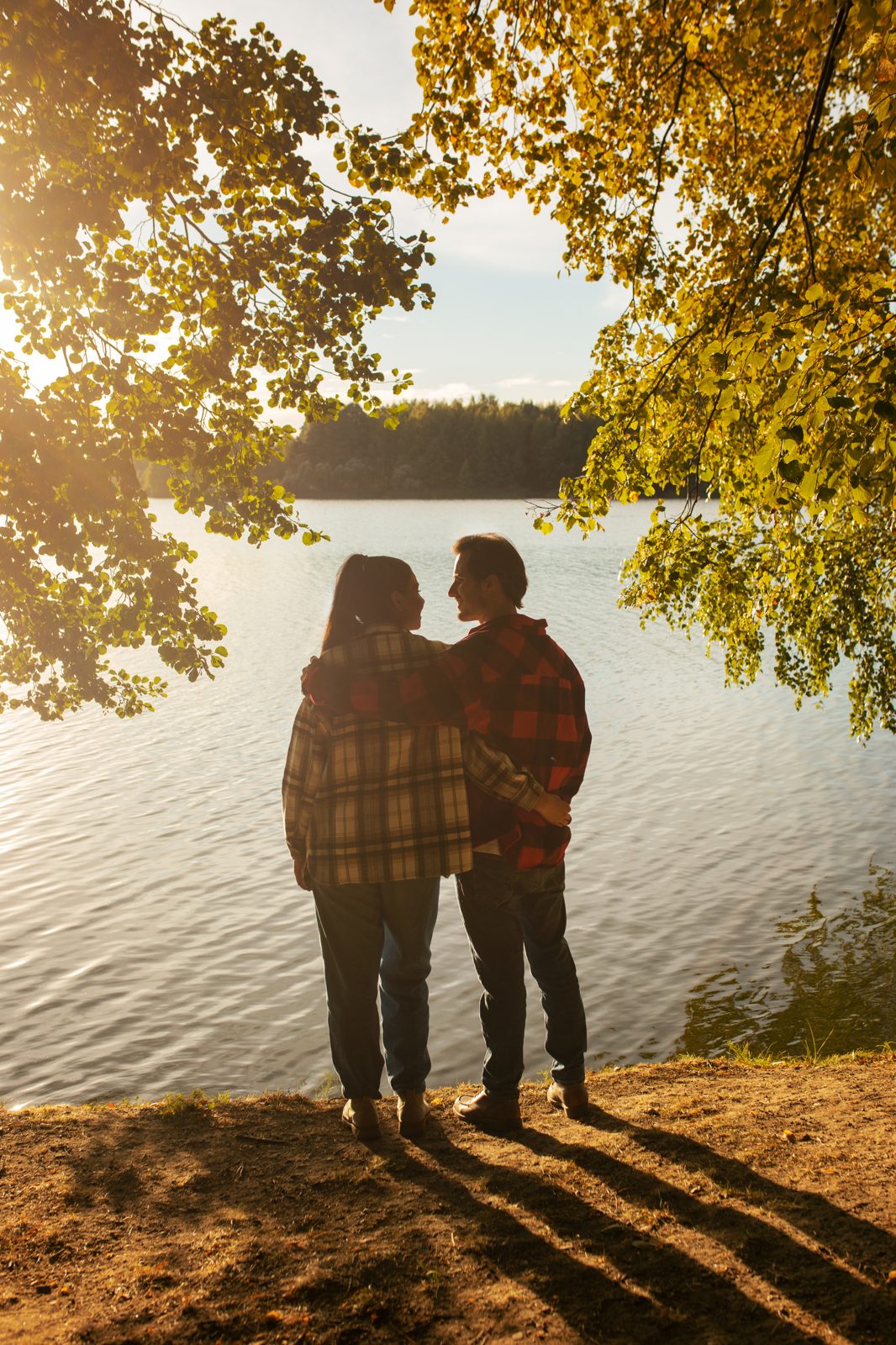 Pareja de espaldas, abrazada a la orilla de un lago en otoño, bajo árboles de hojas amarillas
